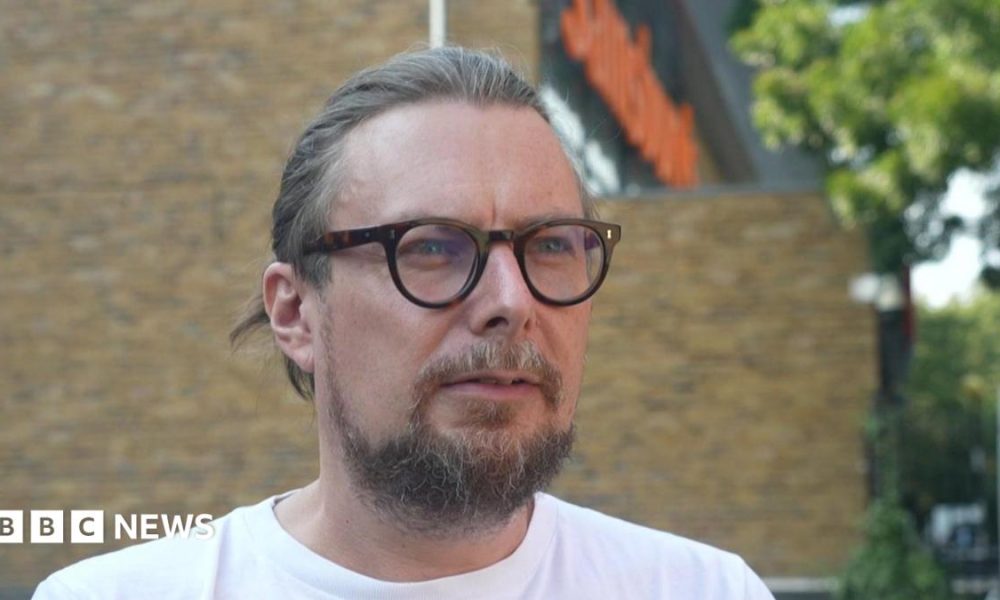 Close-up portrait of Matt Arnold, a man with light brown hair tied back, glasses, and a beard, wearing a white t-shirt outside with a Sainsbury's store blurred in the background.