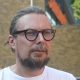 Close-up portrait of Matt Arnold, a man with light brown hair tied back, glasses, and a beard, wearing a white t-shirt outside with a Sainsbury's store blurred in the background.
