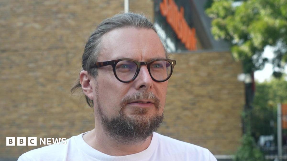 Close-up portrait of Matt Arnold, a man with light brown hair tied back, glasses, and a beard, wearing a white t-shirt outside with a Sainsbury's store blurred in the background.
