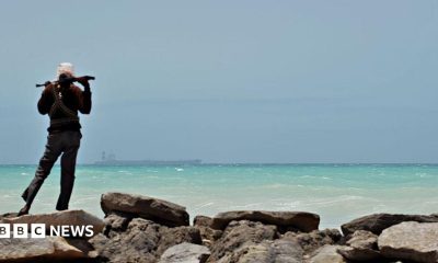 A man - with his back to the camera - stands on some rocks to the left of the picture. Holding a gun across his shoulders, he is looking out to sea where a tanker is in the far distance.