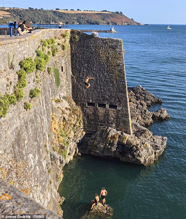 Two young people sit on a jagged rock below a boy as he jumps into the water at Dead Man's Cove in Dartmouth