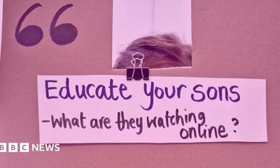 A cardboard sign is held in front of a girls face, her hands are visible on the edges. The phrase 'Rapists - they start as misogynists who aren't challenged' is written on the sign in black marker pen.