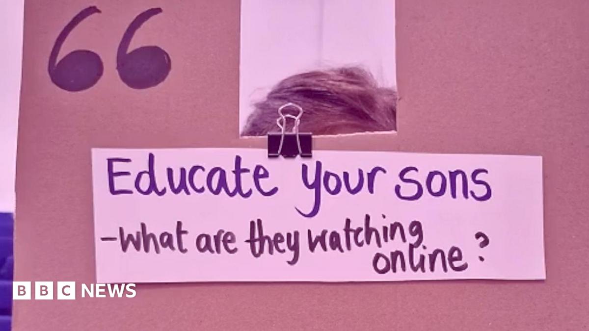 A cardboard sign is held in front of a girls face, her hands are visible on the edges. The phrase 'Rapists - they start as misogynists who aren't challenged' is written on the sign in black marker pen.