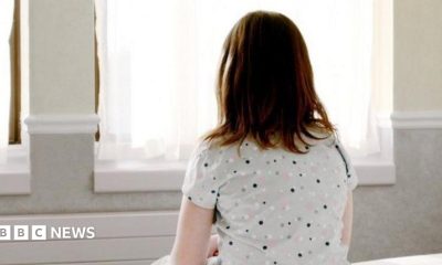 A girl with mid length brown hair sits on a bed facing a window. She is wearing a grey top with black, pink and white dots on it