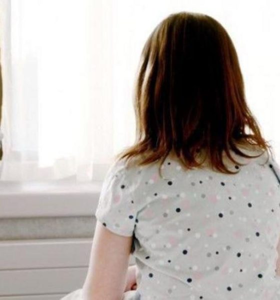 A girl with mid length brown hair sits on a bed facing a window. She is wearing a grey top with black, pink and white dots on it