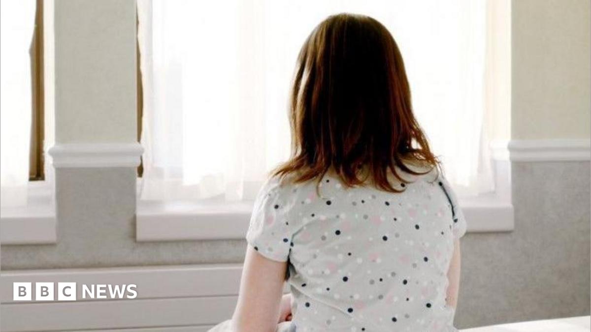 A girl with mid length brown hair sits on a bed facing a window. She is wearing a grey top with black, pink and white dots on it