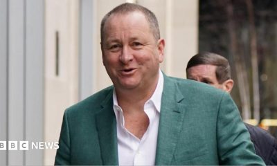 Businessman Mike Ashley wearing a white shirt, and green suit jacket, smiling at the camera as he walks with a man behind him