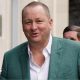 Businessman Mike Ashley wearing a white shirt, and green suit jacket, smiling at the camera as he walks with a man behind him