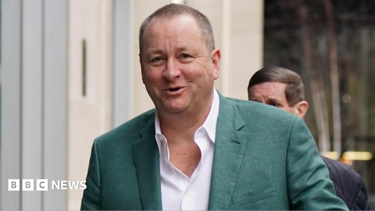 Businessman Mike Ashley wearing a white shirt, and green suit jacket, smiling at the camera as he walks with a man behind him