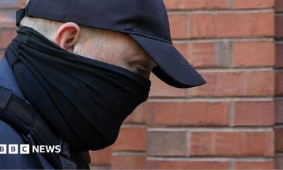 A close-up of the head and shoulders of a man with his face obscured by a black scarf and black cap as he walks towards a red-brick court. His eyes can be seen and his is looking downwards. He is wearing a blue suit jacket and the strap of a bag over his shoulder.