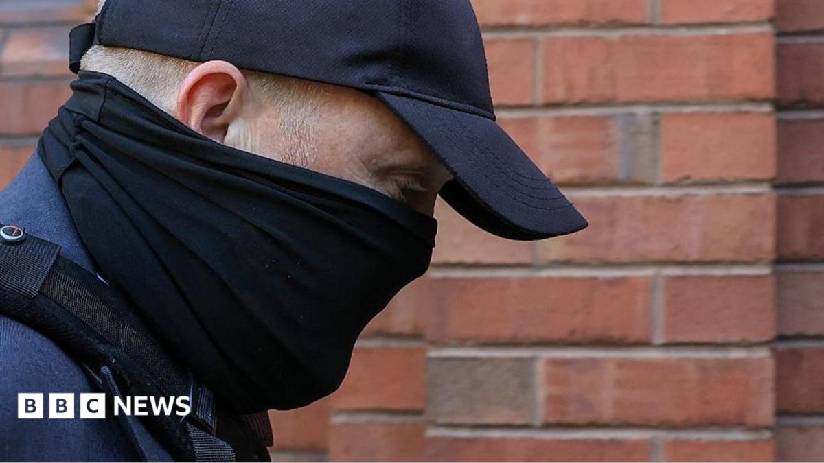 A close-up of the head and shoulders of a man with his face obscured by a black scarf and black cap as he walks towards a red-brick court. His eyes can be seen and his is looking downwards. He is wearing a blue suit jacket and the strap of a bag over his shoulder.