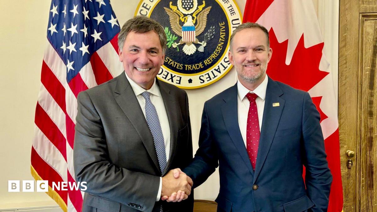 An image showing US trade representative Jamieson Greer (right) shaking hands with Canada-US trade minister Dominic LeBlanc to his left. Behind them are a US and Canada flag with the seal for the US trade representative in the middle. Both men are wearing suits and smiling at the camera.