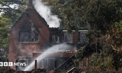 The remains of a burnt out two storey house that still has white puffs of smoke coming away from it. A fire fighter out of shot shoots water up and over the property.