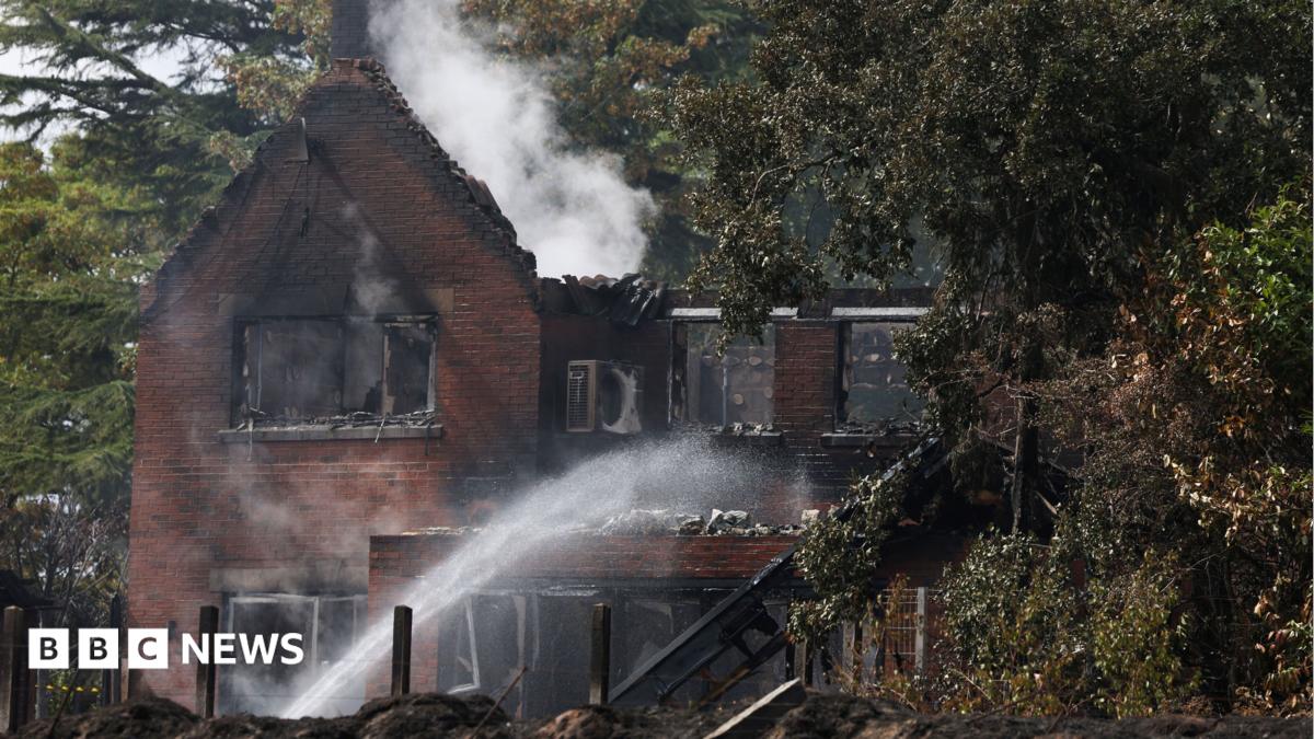 The remains of a burnt out two storey house that still has white puffs of smoke coming away from it. A fire fighter out of shot shoots water up and over the property.