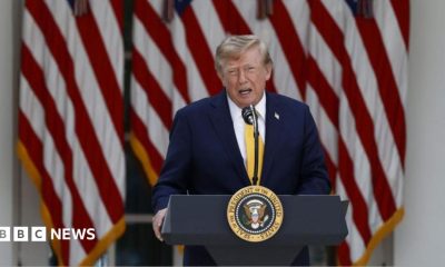 US President Donald Trump delivers remarks outside the White House. He is speaking at a podium with the US presidential seal at the front. He is wearing a dark navy blue suit and a yellow tie. Behind him are several US flags.