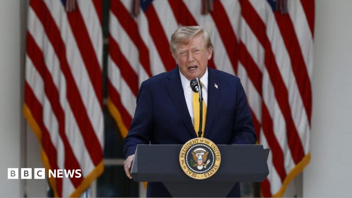 US President Donald Trump delivers remarks outside the White House. He is speaking at a podium with the US presidential seal at the front. He is wearing a dark navy blue suit and a yellow tie. Behind him are several US flags.