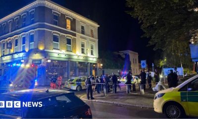 Police officers stand behind a cordon on a residential street in Clapham at night, with forensic tents and emergency vehicles visible near a pub as investigators examine the scene of a fatal shooting.