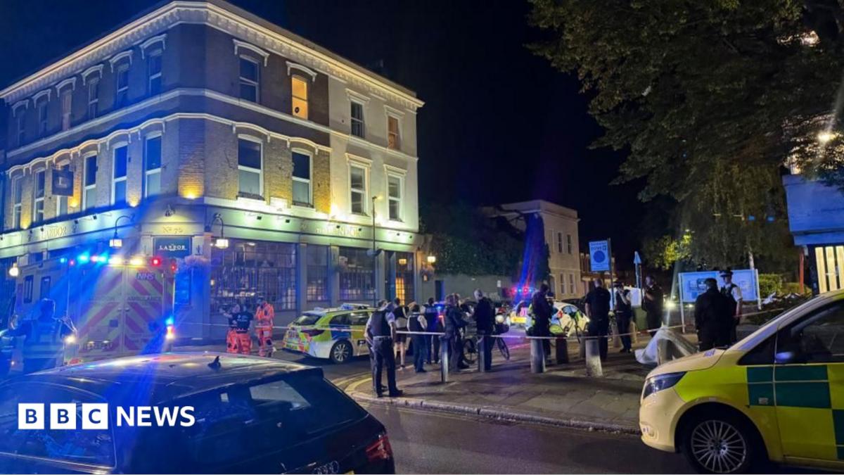 Police officers stand behind a cordon on a residential street in Clapham at night, with forensic tents and emergency vehicles visible near a pub as investigators examine the scene of a fatal shooting.