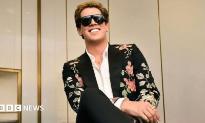 Milo Yiannopoulos wears a colourful suit with dark glasses and a white shirt.