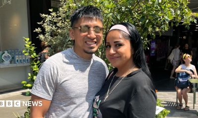 A man and a woman stand together in a sunny plaza. He wears a grey T-shirt and sunglasses. She wears a black T-shirt, necklace and headband