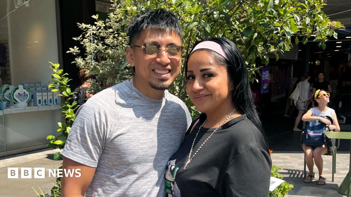 A man and a woman stand together in a sunny plaza. He wears a grey T-shirt and sunglasses. She wears a black T-shirt, necklace and headband