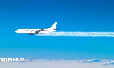 A jet plane flies across the blue sky, with white contrails coming from its engines