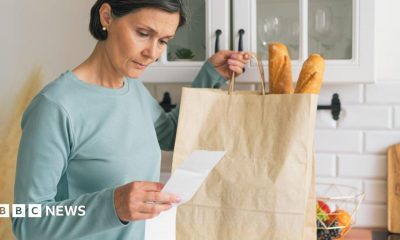 A woman looks at a receipt for her groceries. In her other hand she is holding a paper bag with two baguettes sticking out of the top
