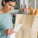 A woman looks at a receipt for her groceries. In her other hand she is holding a paper bag with two baguettes sticking out of the top