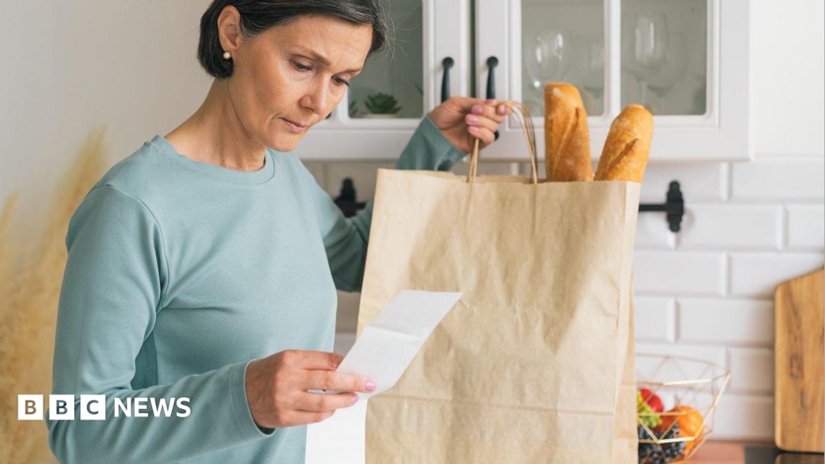 A woman looks at a receipt for her groceries. In her other hand she is holding a paper bag with two baguettes sticking out of the top