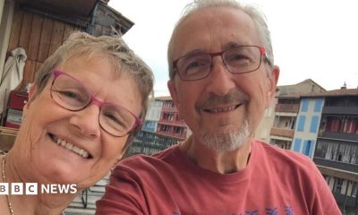 Linda and Geoff Kitchen smile together. Both wear rectangular glasses, with Geoff in a red t-shirt. They appear to be sitting on a balcony, with buildings and the cloudy sky visible in the background.