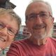 Linda and Geoff Kitchen smile together. Both wear rectangular glasses, with Geoff in a red t-shirt. They appear to be sitting on a balcony, with buildings and the cloudy sky visible in the background.