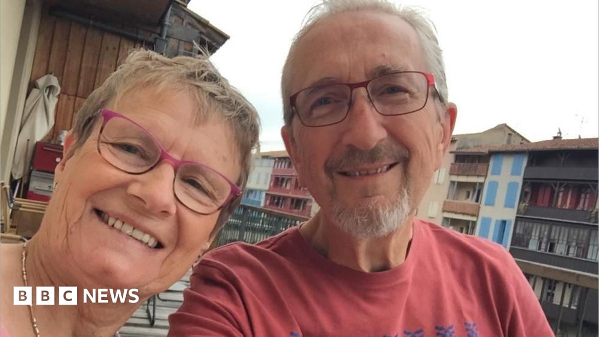 Linda and Geoff Kitchen smile together. Both wear rectangular glasses, with Geoff in a red t-shirt. They appear to be sitting on a balcony, with buildings and the cloudy sky visible in the background.