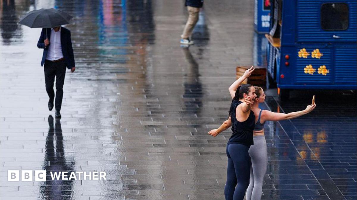 wet street with a man walking along with an umbrella and two women posing for selfie in the rain