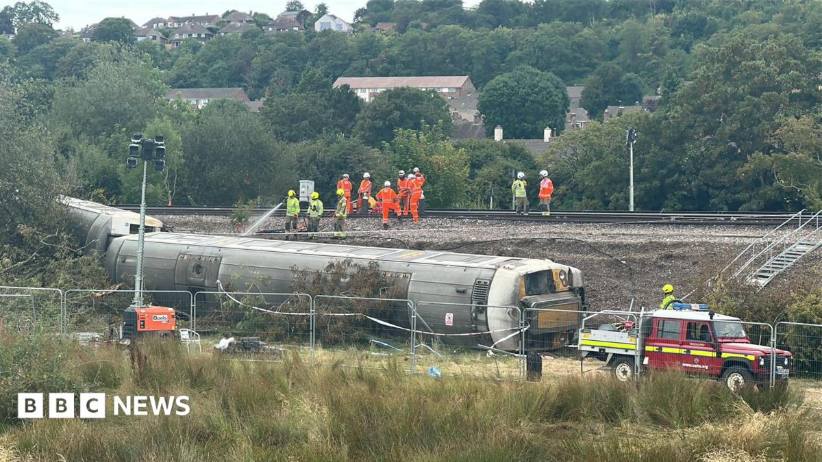 Three derailed train carriages lie at the bottom of a railway embankment near Lewes. Emergency vehicles, fencing and workers in high-visibility clothing can be seen around the site, while crews work on the railway line above.
