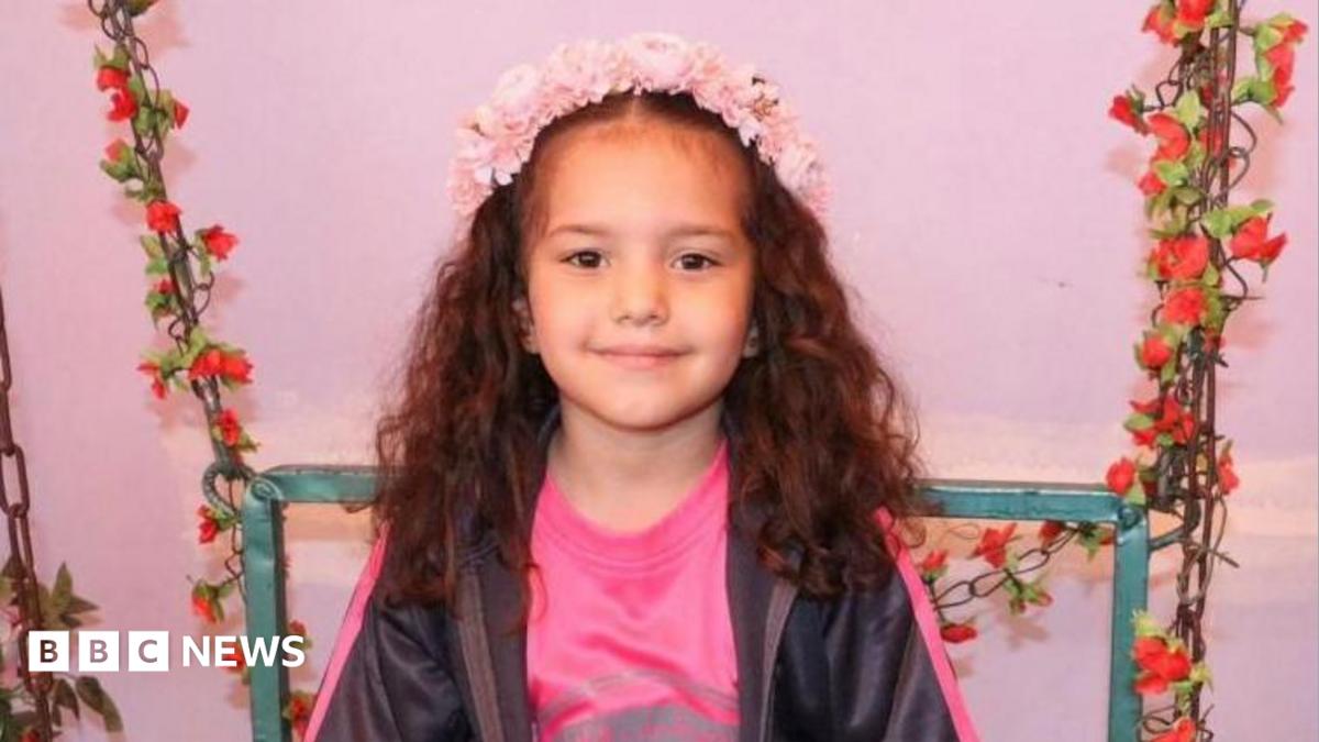 Hind Rajab smiles at the camera. She has long, brown wavy hair and is wearing a pink flower crown on her head. She also has a pink T-shirt and black zip-up on. She is sitting on a chair adorned with garlands of red flowers.