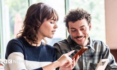 A woman with dark shoulder-length hair points at a smartphone held by a smiling man with curly hair and a moustache.