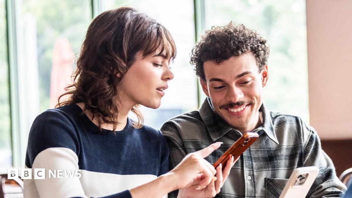 A woman with dark shoulder-length hair points at a smartphone held by a smiling man with curly hair and a moustache.