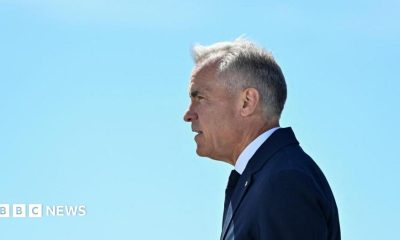 Mark Carney stands in profile against a clear blue sky. His hair is short and grey, and he wears a navy suit and tie and a white short.
