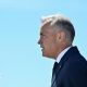 Mark Carney stands in profile against a clear blue sky. His hair is short and grey, and he wears a navy suit and tie and a white short.
