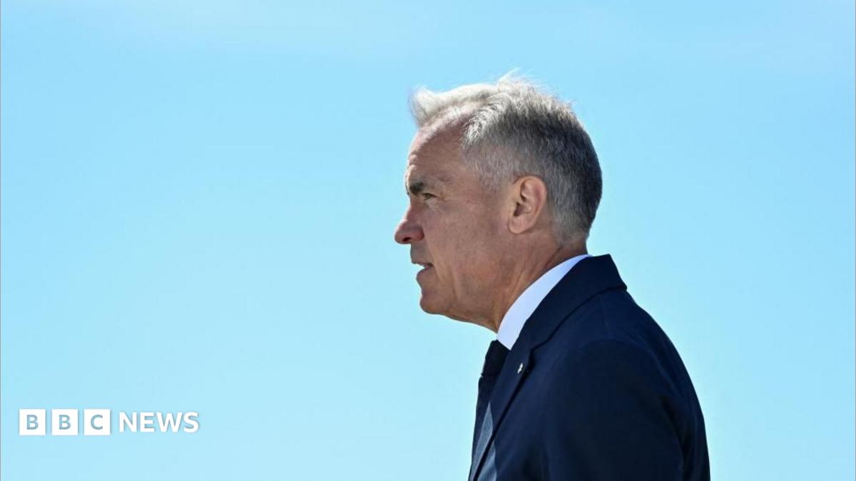 Mark Carney stands in profile against a clear blue sky. His hair is short and grey, and he wears a navy suit and tie and a white short.