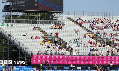 A photo of six women in a race. The floor is blue and the stands are behind which are not full. The signs are pink.