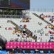 A photo of six women in a race. The floor is blue and the stands are behind which are not full. The signs are pink.