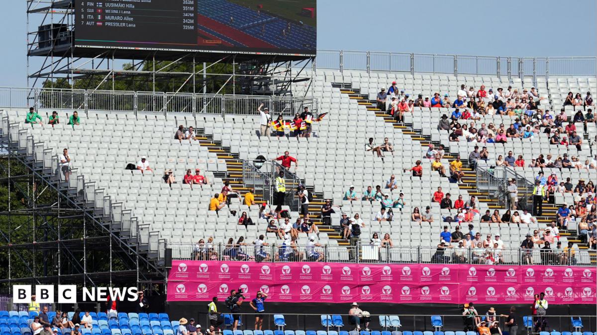 A photo of six women in a race. The floor is blue and the stands are behind which are not full. The signs are pink.