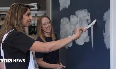Suella Braverman is wearing a dark-coloured top and white dungarees. She is holding a paintbrush and painting white paint on to a blue wall. She is being observed by a woman in the background, who is wearing a black top.