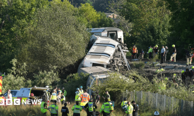 Three train carriages that have derailed. Several emergency workers are congregating by the side of the railway.