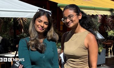 Two women with long dark hair stand in an outdoor square. One wears a turquoise top, the other wears a sleeveless camel top