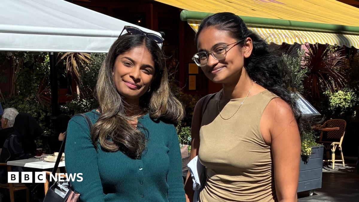 Two women with long dark hair stand in an outdoor square. One wears a turquoise top, the other wears a sleeveless camel top