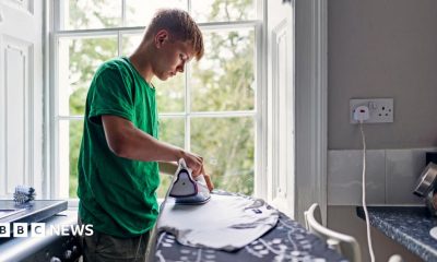 Stock photo shows a person ironing a T shirt in front of a window in their kitchen, next to a plug socket and with the washing machine behind them.