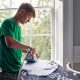 Stock photo shows a person ironing a T shirt in front of a window in their kitchen, next to a plug socket and with the washing machine behind them.