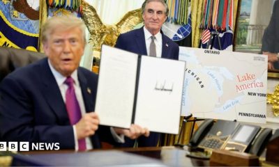 US President Donald Trump holds up a document with his signature in the Oval Office. Begin him is Secretary of the Interior Doug Burghum and a large map showing the US and Canada and the Great Lakes, with Lake Ontario renamed in red script as Lake America.